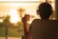 Rear view of woman with headphones and cup of coffee sitting on chair near window and looking on palm trees sea beach at sunrise.