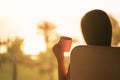 Rear view of woman with headphones and cup of coffee sitting on chair near window and looking on palm trees sea beach at sunrise.