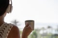 Rear view of woman with headphones and cup of tea looking on palm trees sea beach at sunset. Female relaxing listenning music. Royalty Free Stock Photo