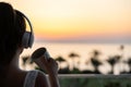 Rear view of woman with headphones and cup of tea looking on palm trees sea beach at sunset. Female relaxing listenning music. Royalty Free Stock Photo