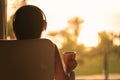 Rear view of woman with headphones and cup of tea sitting on chair near panoramic window and looking on palm trees sea beach