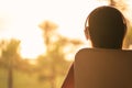 Rear view of woman with headphones sitting on chair near panoramic window and looking on palm trees sea beach at sunset. Female