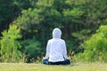 Rear view of woman in hoodie is relaxingly practicing meditation yoga in forest full of grass meadow in summer to attain happiness