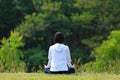 Rear view of woman in hoodie is relaxingly practicing meditation yoga in forest full of grass meadow in summer to attain happiness