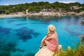 Rear view of a woman sitting on a cliff, admiring the coastal view of Cala Gat beach in Mallorca