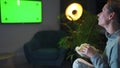 Rear view of a woman sitting on a sofa in the living room in the evening and watching a green TV screen mockup, is