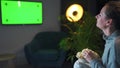 Rear view of a woman sitting on a sofa in the living room in the evening and watching a green TV screen mockup, is