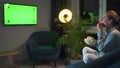 Rear view of a woman sitting on a sofa in the living room in the evening and watching a green TV screen mockup, is