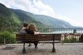 Rear view of a young couple sitting and hugging on a Bench in front of the mountain lake. The man is embracing her girlfriend Royalty Free Stock Photo