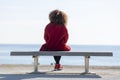 Rear view of a young curly woman wearing red denim jacket sitting on a bench while looking away to horizon over sea Royalty Free Stock Photo