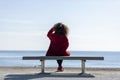 Rear view of a young curly woman wearing red denim jacket sitting on a bench while looking away to horizon over sea Royalty Free Stock Photo