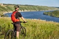 Rear view of the young cyclist stands with mountain bike on the green meadow above big river. Royalty Free Stock Photo