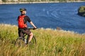 Rear view of the young cyclist stands with mountain bike on the green meadow above big river. Royalty Free Stock Photo