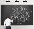 Rear view of young man in formal clothes who is drawing connected business icons on the black chalkboard. The concept of a communi Royalty Free Stock Photo