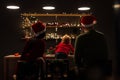 Rear view on young parents and their little child in santa hat. Family sitting at the table in a room with bright garlands. Merry Royalty Free Stock Photo