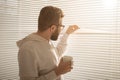 Rear view of young stylish man with beard and coffee peeking through hole in window blinds and looking out into street Royalty Free Stock Photo