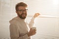 Young stylish man with beard and coffee peeking through hole in window blinds and looking out into street. Concept of Royalty Free Stock Photo