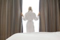 Rear view of young woman in bathrobe opening window curtains at hotel room Royalty Free Stock Photo