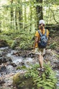 Rear view of young woman hiker in cap with backpack standing on mossy stone near mountain river in forest, walking in nature, eco Royalty Free Stock Photo