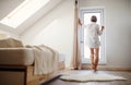 A rear view of young woman with night shirt standing by the window in the morning.