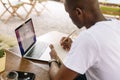 Rearview multicultural dark skin smart man using laptop, taking notes in workbook paper during internship study outdoors Royalty Free Stock Photo