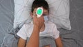 A recovering boy medical protective mask lying in bed and his mother measuring temperature with infrared contactless thermometer,