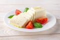 Rectangular slices of mozzarella cheese, fresh basil and quarters of tomato on a white plate over a wooden table. Healhy