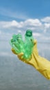 Recycling. Vertical shot of a volunteer wearing yellow rubbish glove holding used plastic bottle against water, river or