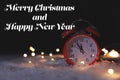 Red alarm clock with fairy lights on snow and message MERRY CHRISTMAS AND HAPPY NEW YEAR against dark background.