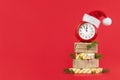 Red alarm clock in Santa Claus hat on wrapped gift boxes decorated fir branches and gold cones. Red background. Concept Royalty Free Stock Photo