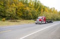 Red icon big rig classic semi truck with flat bed semi trailer running on the autumn road with green and yellow trees Royalty Free Stock Photo