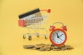 Red analog alarm clock on pile of coins and blurred miniature shopping car with credit card on yellow background Royalty Free Stock Photo