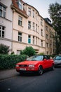A red Audi convertible is parked on the street in front of a few houses. The car has a bright color