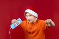 On a red background, a boy in a Santa hat holds a phone in his hand and listens to music through headphones Royalty Free Stock Photo