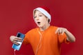 On a red background, a child with a Christmas hat and a yellow T-shirt holds a phone in his hand and listens to music through Royalty Free Stock Photo