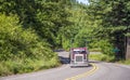 Red big rig classic American semi truck with flat bed semi trailer running on the winding road in green forest Royalty Free Stock Photo