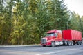 Red big rig low cab semi truck transporting container on the road with green forest on the side