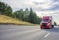 Red big rig semi truck with black grille transporting cargo in semi trailer driving on the straight wide highway road with trees