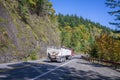 Red big rig semi truck with bulk semi trailer running downhill on the winding autumn road with rock mountain wall and forest