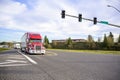 Red big rig semi truck tractor with dry van semi trailer standing on the street crossroad with traffic light starting driving on