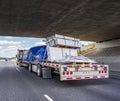 Red big rig semi truck transporting covered and tightened cargo on flat bed semi trailer running under the bridge across wide Royalty Free Stock Photo