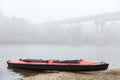 Red and black canoe on lake coast, bridge across river on background, foggy cloudy day, water sport, oar on kayak Royalty Free Stock Photo