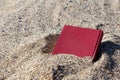 Red book on the sand on a blurry background, covered with sand, buried in the sand.