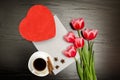 Red box in heart shape, pink tulips, white sheet and a coffee mug. Black table. top view Royalty Free Stock Photo