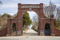 A red brick arch at the entrance of at the Oakland Cemetery with bare winter tree, lush green trees and plants with a blue sky