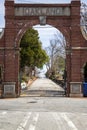 A red brick arch at the entrance of at the Oakland Cemetery with bare winter tree, lush green trees and plants with a blue sky
