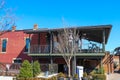 A red brick building with a black metal balcony with tables and stools with bare winter trees in front and lush green plants