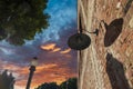 A red brick building with a curved gray light on the wall and tall black lamp posts with tall lush green trees and powerful clouds