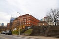 A red brick building surrounded by a staircase, parked cars, tall curved light posts, bare winter trees and skyscrapers