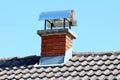 Red brick chimney with shiny metal top protection on new house roof surrounded with dark grey roof tiles and clear blue sky in Royalty Free Stock Photo